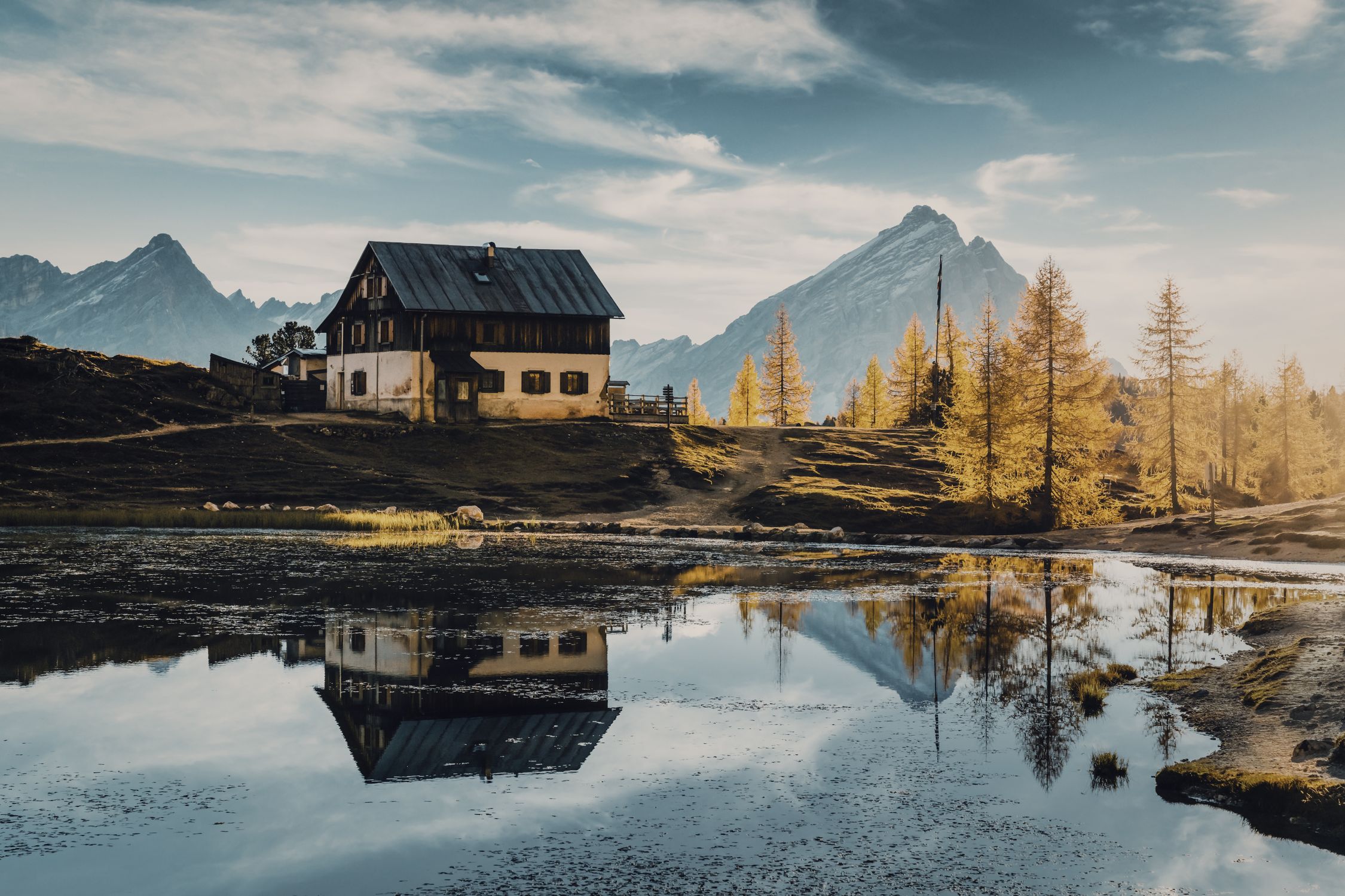 Stone mountain lodge beside a mirror-calm tarn with snow-capped peaks behind